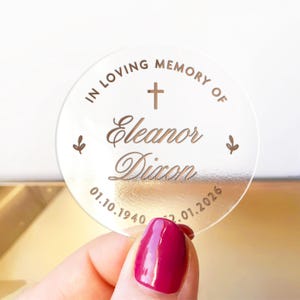 May include: A clear, round memorial keepsake with gold text. The text reads "IN LOVING MEMORY OF Eleanor Dixon" with a cross and dates 01.10.1940 - 12.01.2026. Two small leaf designs flank the text. The keepsake is held by a hand with a magenta nail polish.