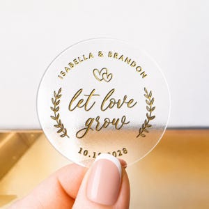 May include: A clear, circular sticker with gold text and design. The text reads "Isabella & Brandon" above two intertwined hearts, with "let love grow" in cursive below. The date "10.1. 028" is at the bottom. Two gold leaf sprigs frame the text.