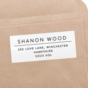 Puede incluir: Una etiqueta de dirección blanca con el nombre "SHANON WOOD" en una fuente limpia y sans-serif. Debajo, la dirección: 205 LOVE LANE, WINCHESTER, HAMPSHIRE, SO23 6QL. La etiqueta está en un sobre marrón claro.