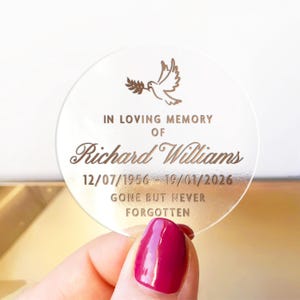 May include: A clear, round memorial keepsake with engraved text and a dove illustration. The text reads "IN LOVING MEMORY OF Richard Williams 12/07/1956 - 19/01/2026 GONE BUT NEVER FORGOTTEN". The keepsake is held by a hand with a red nail polish.