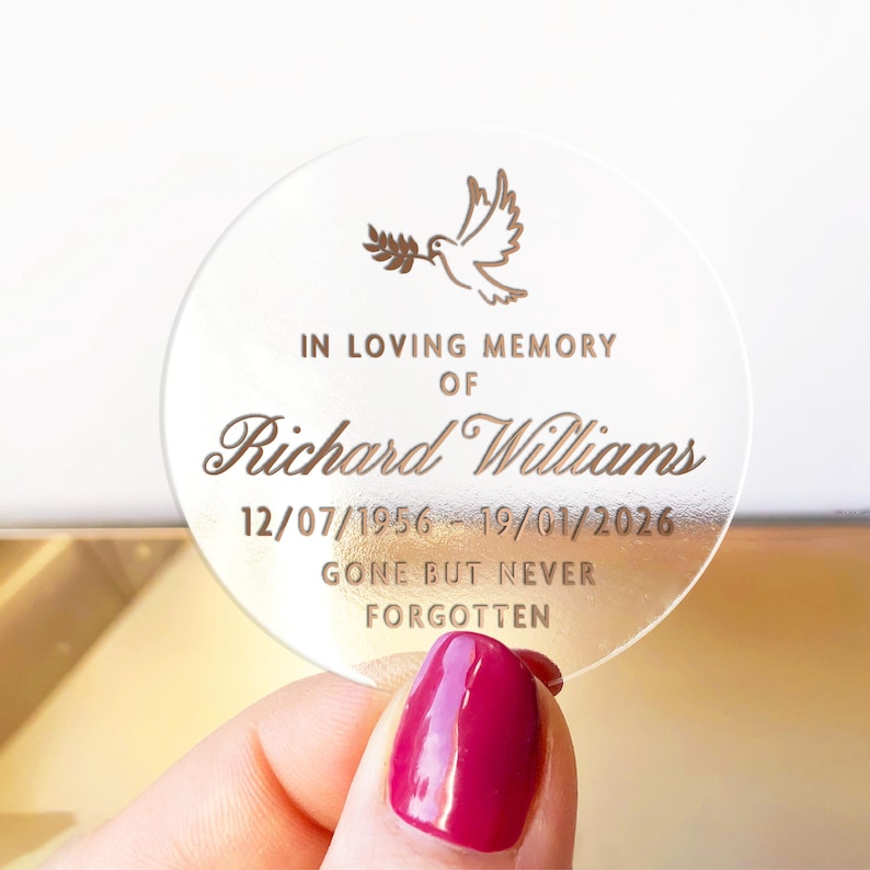 May include: A clear, round memorial keepsake with engraved text and a dove illustration. The text reads "IN LOVING MEMORY OF Richard Williams 12/07/1956 - 19/01/2026 GONE BUT NEVER FORGOTTEN". The keepsake is held by a hand with a red nail polish.