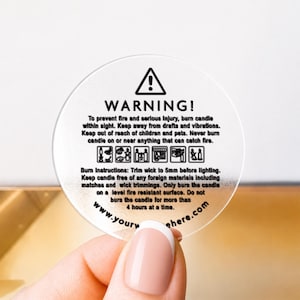 Peut inclure: Un autocollant circulaire transparent avec du texte et des icônes noirs. Il affiche un triangle d'avertissement, des instructions de sécurité pour l'utilisation des bougies et le texte 'WARNING! To prevent fire and serious injury, burn candle'.