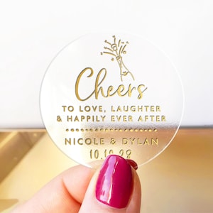 Könnte beinhalten: Ein klarer, runder Aufkleber mit goldfarbenem Schriftzug. Der Text lautet "Cheers" über "To Love, Laughter & Happily Ever After". Darunter stehen "Nicole & Dylan" und das Datum "10.10.28". Oben befindet sich eine Champagnerflaschen-Grafik.