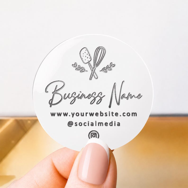 Pu&ograve; includere: Un adesivo circolare bianco con una grafica nera di una spatola e una frusta incrociate con piccoli rametti di foglie. Il testo riporta Business Name, www.yourwebsite.com e @socialmedia con un'icona di Instagram al centro in basso.