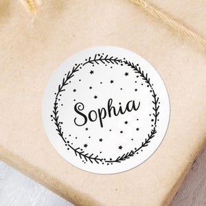 Puede incluir: Una pegatina redonda blanca con un diseño de corona negra y el nombre "Sophia" en letra cursiva. La corona está decorada con pequeñas estrellas.