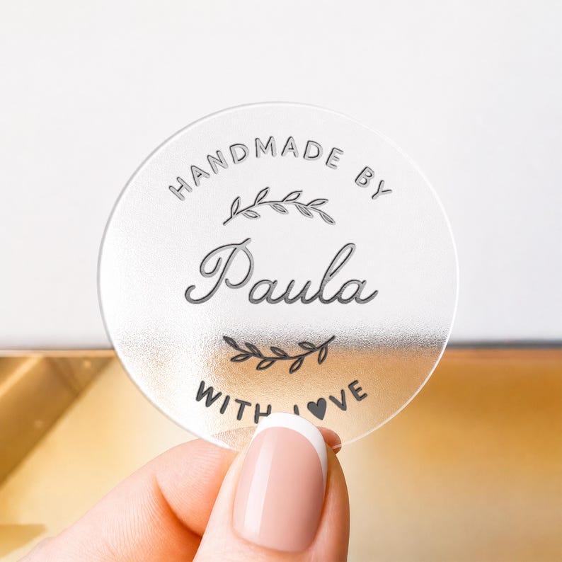 May include: A circular, translucent, frosted acrylic tag featuring the engraved text "HANDMADE BY" at the top, the name "Paula" in a script font in the center, and "WITH LOVE" at the bottom, accented by two small leaf branch illustrations.