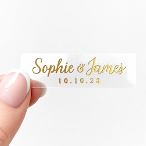 Può includere: Un adesivo rettangolare trasparente con scritte dorate. L'adesivo recita "Sophie & James" sopra la data "10.10.28". L'adesivo è tenuto da una persona con la pelle chiara e una manicure francese.