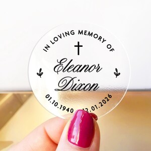 May include: A clear, round memorial sticker with the text "IN LOVING MEMORY OF Eleanor Dixon" and dates 01.10.1940 - 12.01.2026. The design includes a cross and small leaf accents. The sticker is held by a hand with a red nail polish.