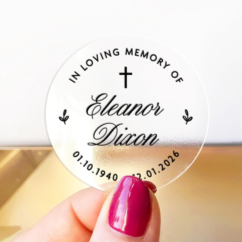 May include: A clear, round memorial sticker with the text "IN LOVING MEMORY OF Eleanor Dixon" and dates 01.10.1940 - 12.01.2026. The design includes a cross and small leaf accents. The sticker is held by a hand with a red nail polish.