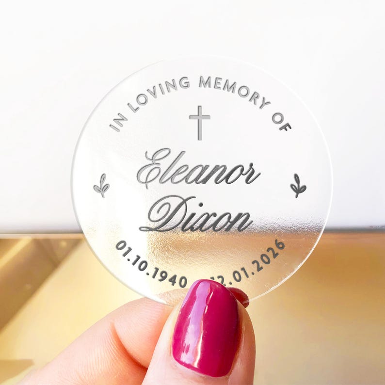 May include: A clear, round memorial keepsake with the inscription "IN LOVING MEMORY OF Eleanor Dixon" and dates 01.10.1940 - 02.01.2026. The design includes a cross and floral accents. The keepsake is held by a hand with a red nail polish.