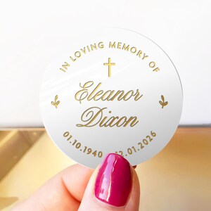 May include: A white, round memorial sticker with gold text. The text reads "IN LOVING MEMORY OF Eleanor Dixon" with a cross and dates 01.10.1940 - 22.01.2026. Two small leaf designs are on the sides.
