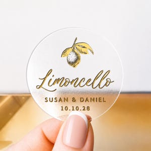 May include: A clear, circular sticker featuring gold lettering and a lemon design. The word "Limoncello" is written in a decorative font, with "SUSAN & DANIEL" and the date "10.10.28" below. The sticker is held by a hand.