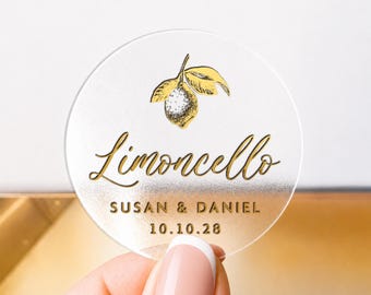 May include: Clear round sticker with gold text and a lemon illustration. The word "Limoncello" is written in elegant script, with "SUSAN & DANIEL" and the date "10.10.28" below. The sticker is held by a hand.