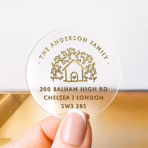 May include: A circular, frosted acrylic sticker featuring gold foil text and graphics. The design includes a house icon surrounded by leaves, the text THE ANDERSON FAMILY, and the address 200 BALHAM HIGH RD, CHELSEA | LONDON, SW3 2BS.