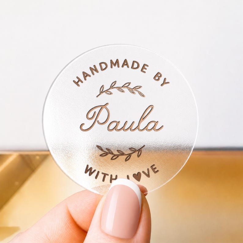May include: A circular, translucent acrylic tag featuring the engraved text "HANDMADE BY Paula WITH LOVE" in a dark brown font. The design includes two decorative leaf sprigs positioned above and below the name, with a small heart icon.