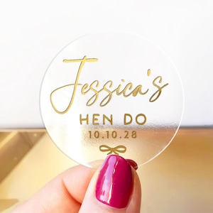 Pode incluir: Um autocolante redondo transparente com letras douradas que diz "Jessica's HEN DO 10.10.28" e um pequeno laço. O autocolante é segurado por uma mão com manicure rosa.