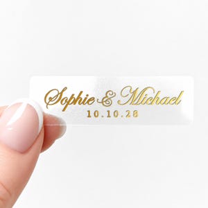 May include: A clear rectangular sticker with gold lettering. The text reads "Sophie & Michael" above the date "10.10.28". The sticker is held between two fingers.