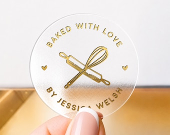 Könnte beinhalten: Ein kreisförmiges, mattiertes, halbtransparentes Acrylschild mit goldener Schrift BAKED WITH LOVE und BY JESSICA WELSH, einer zentralen Grafik aus einem gekreuzten Nudelholz und Schneebesen sowie zwei kleinen goldenen Herzsymbolen.