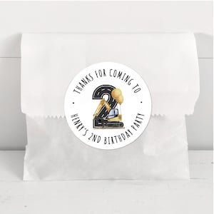 May include: A white paper bag with a round sticker. The sticker says "Thanks for coming to Henry's 2nd Birthday Party" and features a black number 2, a yellow excavator, and two yellow balloons.