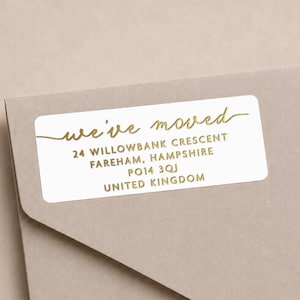 Puede incluir: Una etiqueta de envío rectangular blanca con esquinas redondeadas que presenta texto en lámina dorada que dice we've moved seguido de la dirección 24 Willowbank Crescent, Fareham, Hampshire, PO14 3QJ, United Kingdom, sobre un sobre marrón claro.