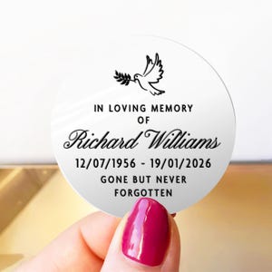 May include: A round, white memorial sticker with black text and a dove illustration. The text reads "IN LOVING MEMORY OF Richard Williams 12/07/1956 - 19/01/2026 GONE BUT NEVER FORGOTTEN". The sticker is held by a hand with a red nail polish.
