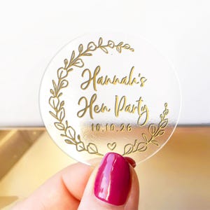 May include: Clear, round sticker with gold lettering and a gold floral wreath. The text reads "Hannah's Hen Party" and "10.10.26". A small heart is below the date. The sticker is held by a hand with a magenta nail polish.