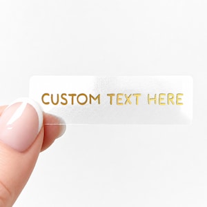 Può includere: Un adesivo rettangolare trasparente con la scritta dorata "CUSTOM TEXT HERE". L'adesivo è tenuto dalle dita di una persona su uno sfondo bianco. L'adesivo è leggermente riflettente.