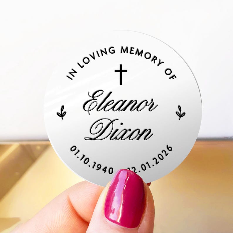May include: A white, round memorial sticker with black text. The text reads "IN LOVING MEMORY OF" above a cross, followed by "Eleanor Dixon" and the dates 01.10.1940 - 22.01.2026. Small leaf designs flank the text.