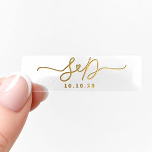 May include: A clear rectangular sticker with gold script lettering. The script reads "S&D" with a heart symbol between the letters, and the date "10.10.28" below. The sticker is held by a person's hand.