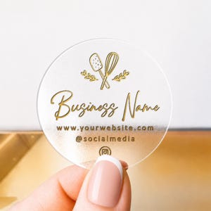 Pu&ograve; includere: Un adesivo circolare, satinato e semitrasparente con dettagli in lamina d'oro. Il design include un'icona di una frusta e una spatola, il testo "Business Name", "www.yourwebsite.com", "@socialmedia" e un'icona Instagram al centro in basso.