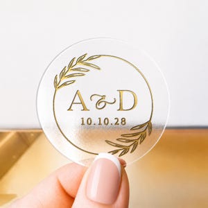 Könnte beinhalten: Ein klarer, runder Aufkleber mit goldfarbenen Buchstaben und einem goldenen Blattdesign. Der Aufkleber zeigt die Initialen "A & D" und das Datum "10.10.28". Der Aufkleber wird von einer Person mit French Manicure gehalten.