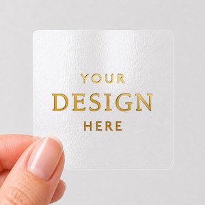 May include: A square, transparent sticker featuring the gold-coloured text YOUR DESIGN HERE printed in the centre. The sticker has a smooth, semi-gloss finish and rounded corners, providing a clear surface for custom branding or labelling applications.