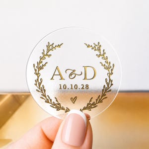 Op de afbeelding: Een transparante, ronde sticker met gouden letters en een lauwerkransontwerp. De tekst luidt "A & D" met de datum "10.10.28" en een hartsymbool. De sticker wordt vastgehouden door een hand met een French manicure.
