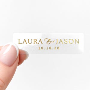 Może przedstawiać: Przezroczysta prostokątna naklejka ze złotym napisem "LAURA & JASON" nad datą "10.10.28". Naklejka jest trzymana między dwoma palcami na białym tle.