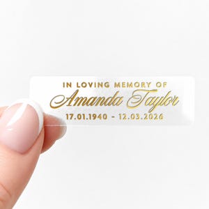 May include: A clear rectangular sticker with gold text. The text reads "IN LOVING MEMORY OF Amanda Taylor 17.01.1940 - 12.03.2026". The sticker is held by a person with a light skin tone.