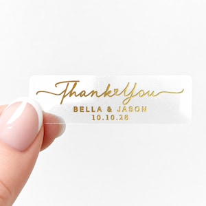 Puede incluir: Una pegatina rectangular transparente con texto dorado que dice "Thank You" en una fuente cursiva. Debajo de la escritura está "BELLA & JASON" y la fecha "10.10.28". La pegatina está sujeta por una mano.
