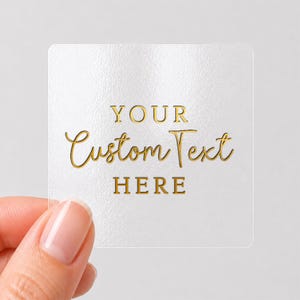 May include: A square, transparent adhesive sticker featuring gold foil lettering. The text is arranged in three lines, with the words YOUR and HERE in a simple serif font, and the words Custom Text in a cursive script font in the center.