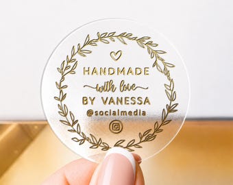 May include: A circular, frosted acrylic tag featuring a gold-colored laurel wreath design. The text reads "HANDMADE with love BY VANESSA @socialmedia" with a small heart icon above the text and an Instagram logo icon at the bottom center.