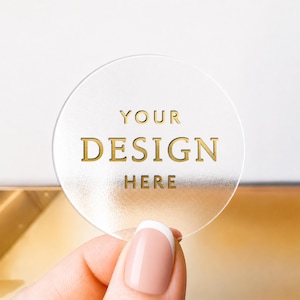 May include: A circular, frosted, semi-transparent acrylic disc featuring the gold-coloured text "YOUR DESIGN HERE" printed in the centre. The disc has a smooth, matte-like texture and is held up against a plain, light-coloured background.