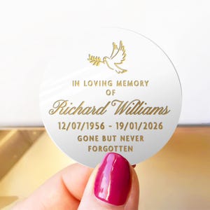 May include: A white, round memorial keepsake with gold text and a dove illustration. The text reads "IN LOVING MEMORY OF Richard Williams 12/07/1956 - 19/01/2026 GONE BUT NEVER FORGOTTEN."