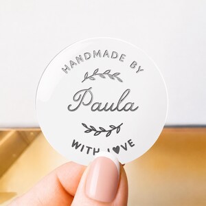 May include: A circular white sticker featuring the text HANDMADE BY Paula WITH LOVE in dark gray lettering. The design includes a decorative leaf branch graphic above the name and a smaller leaf branch graphic below the name Paula.