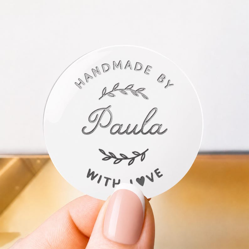 May include: A circular white sticker featuring the text HANDMADE BY Paula WITH LOVE in dark gray lettering. The design includes a decorative leaf branch graphic above the name and a smaller leaf branch graphic below the name Paula.