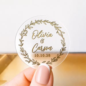 May include: A clear, circular sticker featuring gold lettering and a gold leaf wreath design. The sticker displays the names "Olivia & Carson" and the date "10.10.28". The sticker is held by a hand with a French manicure.