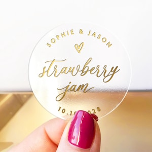Op de afbeelding: Een heldere, ronde sticker met gouden letters. Er staat "SOPHIE & JASON" boven "Strawberry Jam" en de datum "10.10.2028". Een gouden hart staat tussen de namen. De sticker wordt vastgehouden door een hand met magenta nagellak.