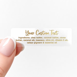 May include: A clear rectangular label with gold text that reads "Your Custom Text." Below, the ingredients are listed: shea butter, coconut butter, cocoa butter, coconut oil, beeswax, olive oil, vitamin E oil, color pigment & essential oil.