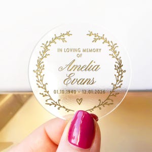 May include: A clear, round memorial sticker with gold text and a gold laurel wreath design. The text reads "IN LOVING MEMORY OF Amelia Evans 01.10.1940 - 12.01.2026" with a heart symbol. The sticker is held by a person with a red nail polish.