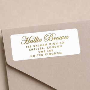 May include: A rectangular white address label with rounded corners featuring gold foil text that reads Hallie Brown, 198 Balham High Rd, Chelsea, London, SW3 2BS, United Kingdom. The label is applied to the corner of a light brown envelope.