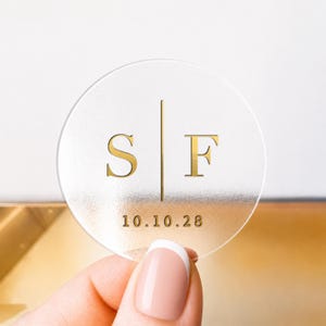 May include: A clear, circular sticker with gold lettering. It displays the letters "S" and "F" divided by a vertical line, with the date "10.10.28" beneath. The sticker is held by a person with a French manicure.