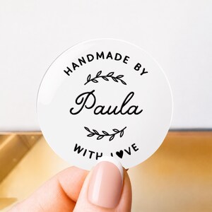 May include: A circular white sticker featuring black text that reads HANDMADE BY Paula WITH LOVE. The design includes a decorative leafy branch graphic above and below the name Paula, with a small heart replacing the letter O in LOVE.