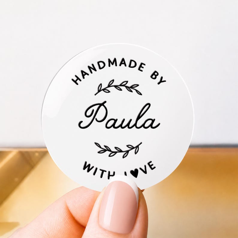 May include: A circular white sticker featuring black text that reads HANDMADE BY Paula WITH LOVE. The design includes a decorative leafy branch graphic above and below the name Paula, with a small heart replacing the letter O in LOVE.